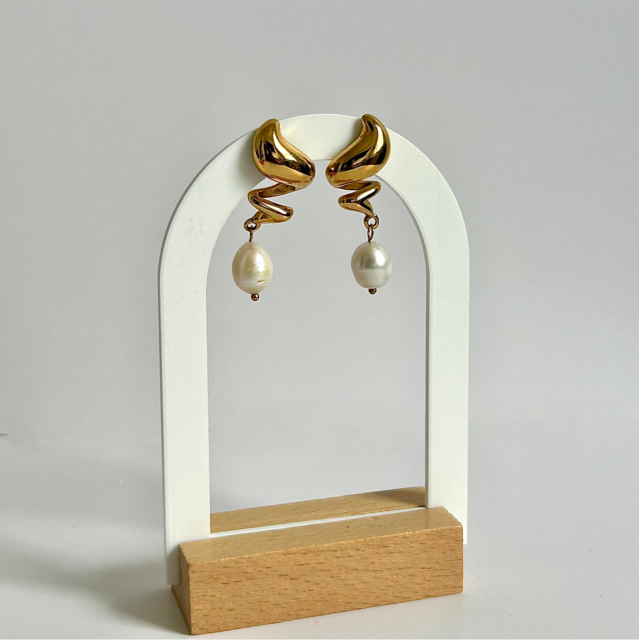Gold spiral earrings with pearl drops on a white arch stand against a light gray background
